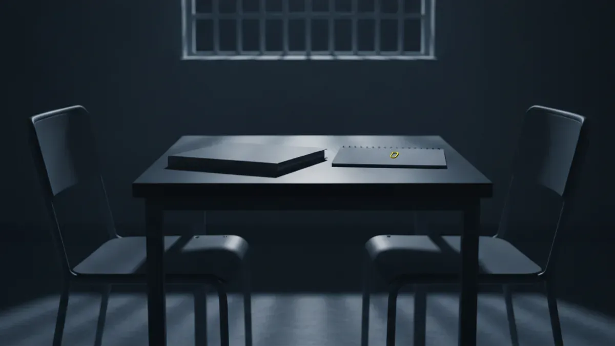 Sala de entrevistas de un centro penitenciario en penumbra, con dos sillas vacías enfrentadas, una carpeta gris y una libreta con un clip amarillo sobre la mesa, y una ventana con barrotes al fondo